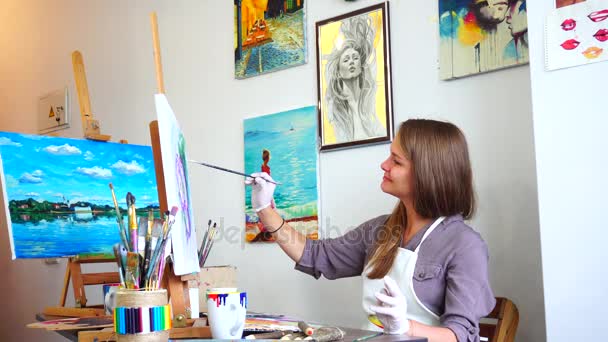 Girl Artist Sits in Profile to Camera and Looking at Picture, Draws Brushstrokes and Lines on Canvas With Brush at Easel in Bright Art Class Studio. - Footage, Video