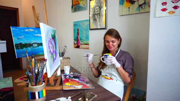 Joven artista mujer toma taza en las manos y beber, sentado con cepillo en la mano cerca de cuadro completo en caballete en silla en la mesa en el estudio
. - Metraje, vídeo