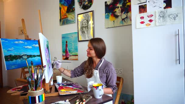 Ženy sedí umělec v profilu do kamery poklesy štětec v programu Malování a způsobují tahy na plátno s kartáčem, sedí Onchair na stojan ve výtvarce. - Záběry, video