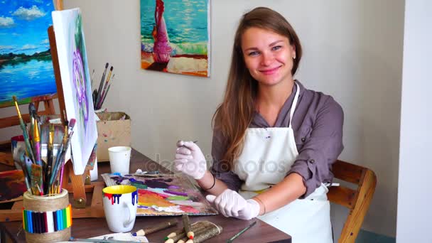 Beautiful Girl Artist Looks at Camera and Smiling While Holding Brush in Hand and Draws Picture at Easel Sitting on Chair in Art Studio. - Footage, Video