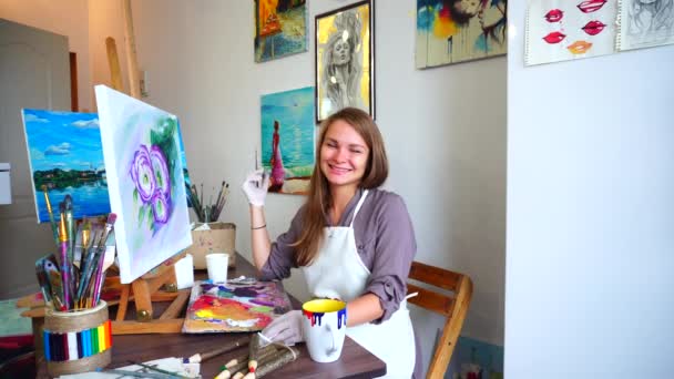 Portret artysty kobieta uśmiecha się i śmieje się, patrząc w obiektyw fotografa i siedząc przy sztalugach w Art Studio. - Materiał filmowy, wideo