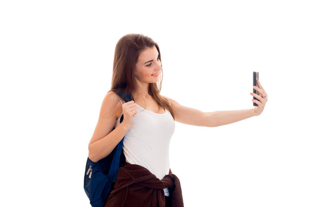 cheerful young brunette student girl with blue backpackand and mobile phone in her hands makes selfie isolated on white background - Foto, afbeelding