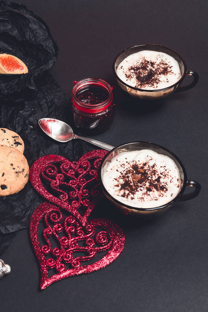 Dos tazas de café, capuchino cerca de corazones rojos sobre fondo de mesa negro. San Valentín. Amor. Desayuno romántico
.  - Foto, Imagen