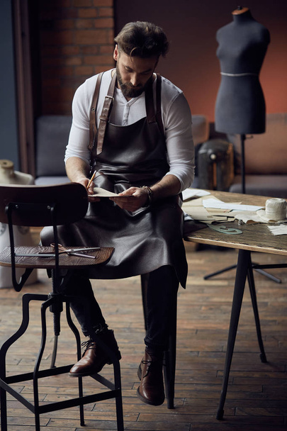 Young handsome tailor with beard in white shirt and leather apron sitting on wooden table with threads and drawing in amazing atelier with antique furniture - Valokuva, kuva