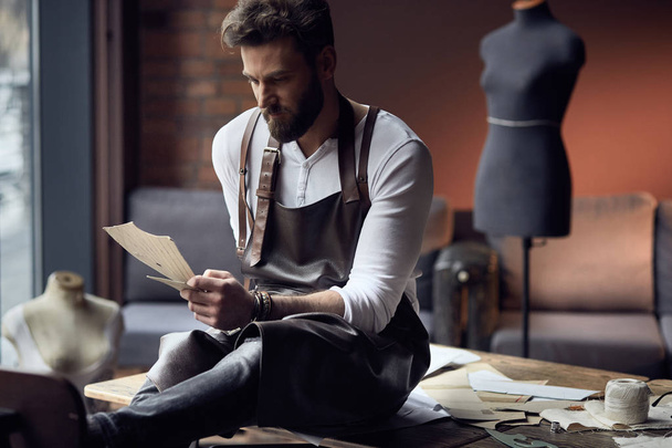 Young handsome tailor with beard in white shirt and leather apron sitting on wooden table with threads in amazing atelier with antique furniture and mannequin on background - Zdjęcie, obraz