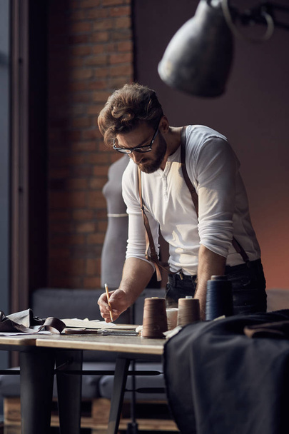 Young serious tailor with beard and glasses in white shirt with leather suspenders working near wooden table with threads in amazing atelier with antique furniture and mannequin on background - Фото, изображение