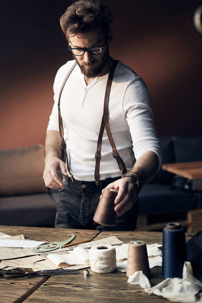 Young cute tailor with beard and glasses in white shirt with leather suspenders working near wooden table with threads in amazing atelier with antique furniture and mannequin on background - Foto, immagini