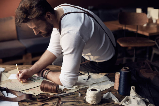 Handsome tailor male with beard in white shirt with brown leather suspenders working near wooden table with threads in amazing atelier with antique furniture and mannequin on background - Zdjęcie, obraz