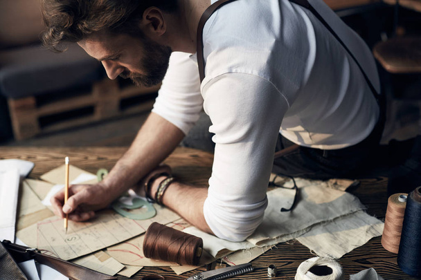 Handsome tailor male with beard in white shirt with brown leather suspenders drawing near wooden table with threads and scissors in amazing atelier with antique furniture and mannequin on background - Foto, Imagem