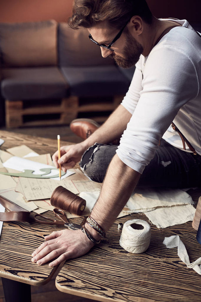 Handsome tailor male with beard and glasses in white shirt with brown leather suspenders drawing near wooden table with threads and scissors in amazing atelier with antique furniture  - Foto, Bild