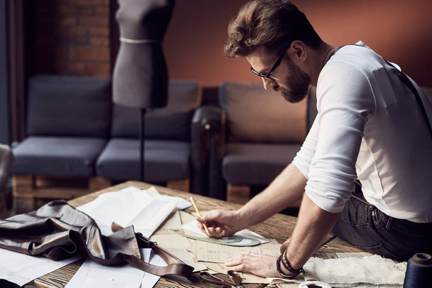 Handsome tailor male with beard and glasses in white shirt with brown leather suspenders drawing near wooden table with threads, apron and scissors in amazing atelier with antique furniture and mannequin on background - Foto, imagen
