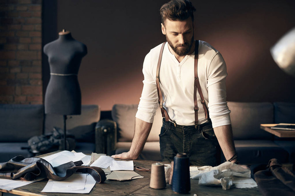 Beautiful serious tailor with beard in white shirt with brown leather suspenders standing near wooden table with threads, apron and scissors and thinking in amazing atelier with antique furniture  - Foto, Bild