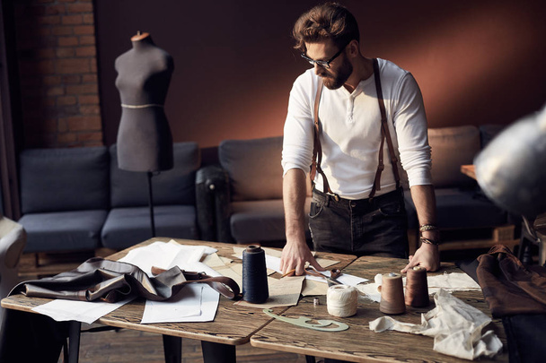 Handsome serious tailor with beard and glasses in white shirt with brown leather suspenders working near wooden table with threads, apron and scissors in amazing atelier with antique furniture and mannequin on background - Foto, afbeelding