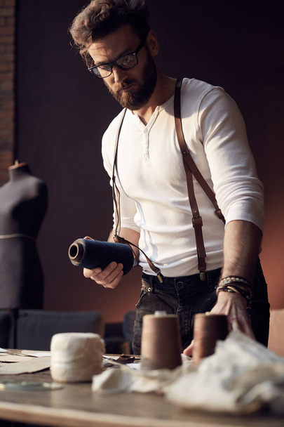 Great serious tailor with beard and glasses in white shirt with brown leather suspenders working near wooden table with threads and scissors in amazing atelier with antique furniture and mannequin on background - Valokuva, kuva