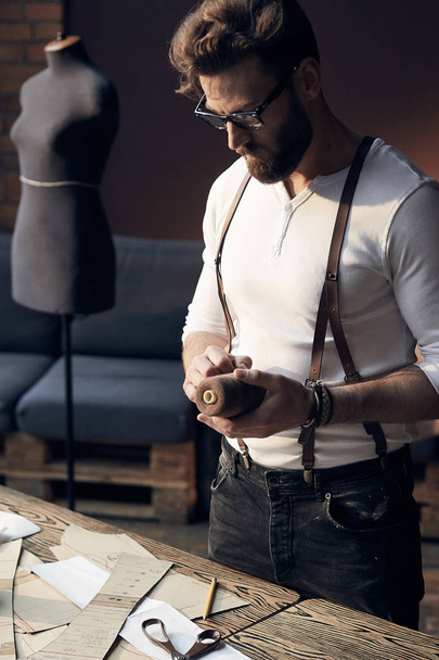 Cute serious tailor with beard and glasses in white shirt with brown leather suspenders working near wooden table with threads and tailoring equipment in amazing atelier with antique furniture and mannequin on background - Foto, Bild