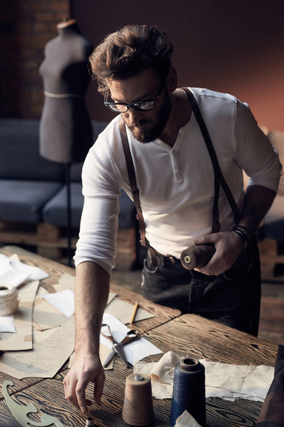 Young serious tailor with beard and glasses in white shirt with brown leather suspenders working near wooden table with threads and tailoring equipment in amazing atelier with antique furniture and mannequin on background - Foto, imagen