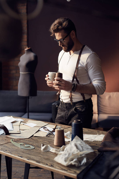 Serious tailor with beard and glasses in white shirt with brown leather suspenders working near wooden table with threads and tailoring equipment in amazing atelier with antique furniture and mannequin on background - Zdjęcie, obraz