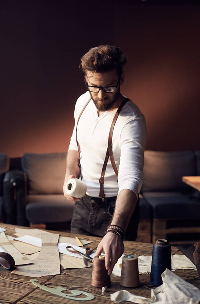 Serious tailor with beard and glasses in white shirt with brown leather suspenders working near wooden table with threads and tailoring equipment in amazing atelier with antique furniture  - Foto, Bild