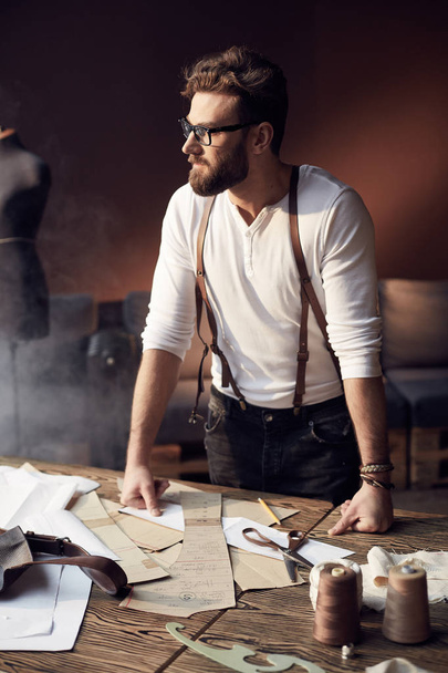 Serious young tailor with beard and glasses in white shirt with brown leather suspenders standing near wooden table with threads and tailoring equipment and looking at the window in amazing atelier with antique furniture - Fotó, kép