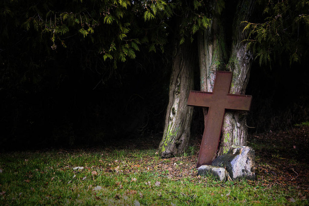 Rostiges Kreuz aus Metall lehnt im Schatten an alten Baumstämmen - Foto, Bild