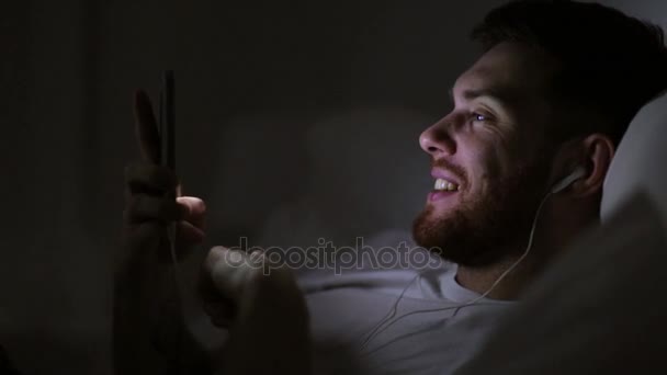 man with smartphone and earphones in bed at night - Footage, Video