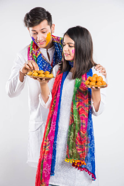 νεαρό ζευγάρι Ινδιάνων με παραδοσιακή ενδυμασία με ζωγραφισμένα πρόσωπα που κρατούν πιάτα γεμάτα χρώματα ή γλυκά που γιορτάζουν το Holi, απομονωμένα σε λευκό φόντο - Φωτογραφία, εικόνα