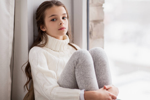 sad girl sitting on sill at home window in winter - Foto, Imagen