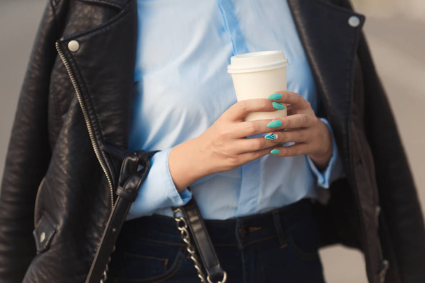 Stylish woman with colorful manicure in leather jacket holding in hands a paper cup of coffee  - Fotoğraf, Görsel