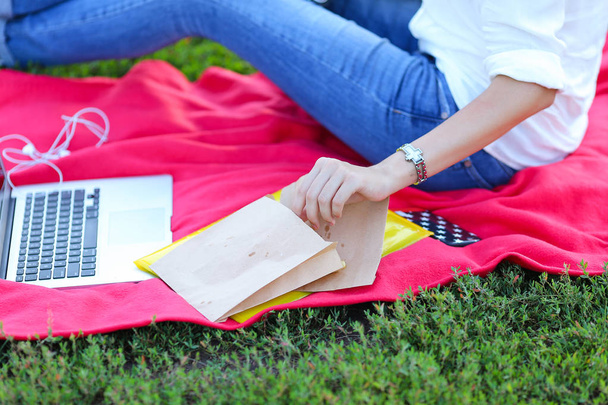Nuori tyttö istuu nurmikolla puistossa ja työskentelee kannettavan tietokoneen ja syö pikaruokaa
 - Valokuva, kuva