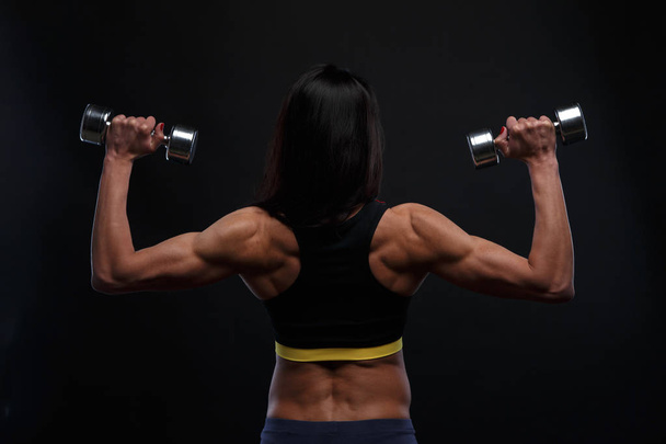 Back of beautiful athletic girl working with weights on a dark background - Photo, Image