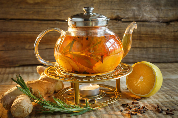 Glass tea pot with hot tasty tea with lemon, herbs,. Yellow lemon and tea. Tea pot on warm colors on wooden background. Herbal tea in a glass tea pot. - Photo, Image
