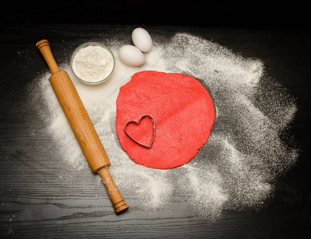 Koło czerwonego ciasta z wycięcie w kształcie serca. Czarny stół posypany, wałkiem i jajka. Widok z góry - Zdjęcie, obraz