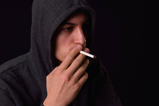 Teenager in a black hooded sweatshirt smoking a cigarette on a d - Valokuva, kuva