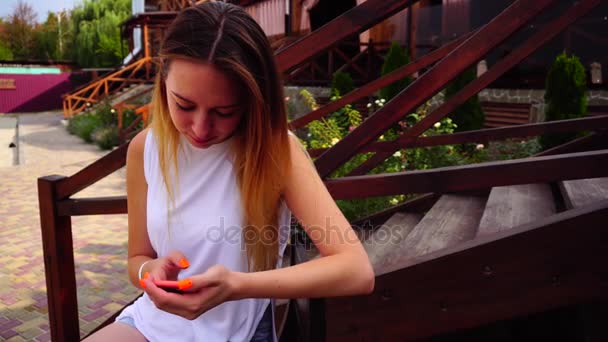 Hezká mladá dívka drží telefon v ruce, výměnu zpráv, čte Sms a usmívá se, sedí na lavičce na pozadí záhonů a venkovní schody . - Záběry, video