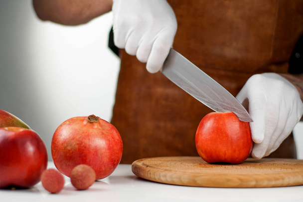 Chef in white gloves cutting ripe apple on a wooden cutting board near other fruits. Healthy lifestyle concept - Fotó, kép