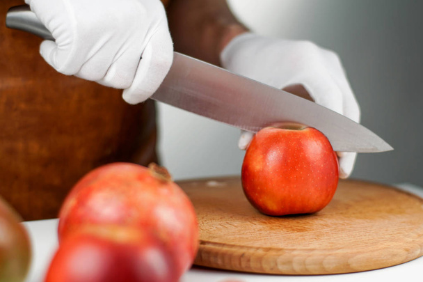 Close-up on hands in white gloves cutting an apple with the help of big sharp knife - Fotó, kép