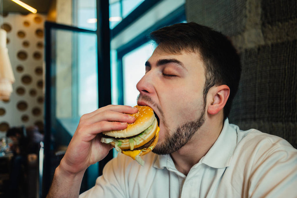 молодий чоловік з бородою їсть бургер в ресторані і насолоджується смаком
 - Фото, зображення