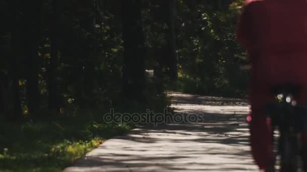 Adam kırmızı spor kıyafetli bir ormandaki bir asfalt yol boyunca bir bisiklet ile gidiyor - Video, Çekim