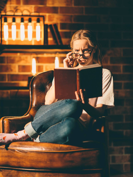 Dziewczyna w przytulnym ciemnym pokoju, czytająca książkę w okularach. Blond i ciepłe światło lampy - Zdjęcie, obraz