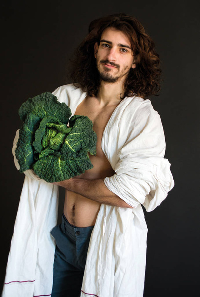 photo guy with long curly hair in a white drapery holding cabbage in hands - Photo, Image