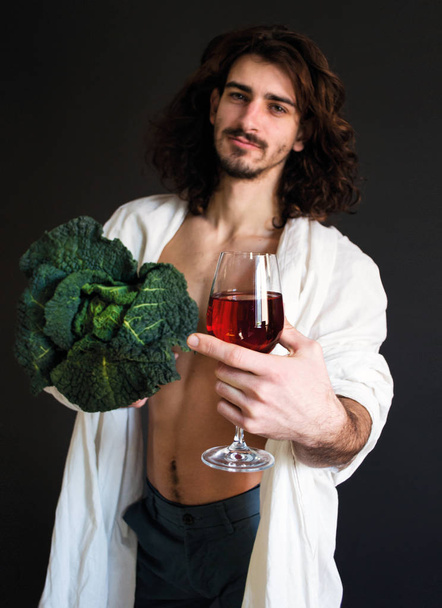 photo beautiful guy in a white drapery holding fresh green cabbage and a glass of wine in hand - Фото, изображение