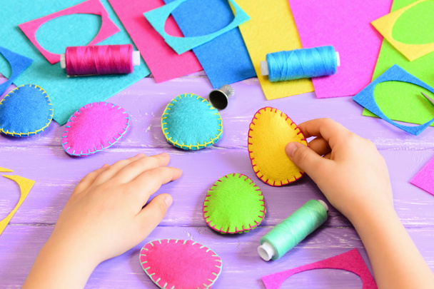 Child hold a felt Easter egg in his hand. Small child made colourful felt Easter crafts diy. Tools and materials for sewing handicraft on wooden table. Easy fun kids Easter crafts activities concept - Photo, Image
