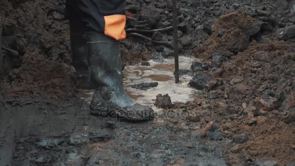 Hombre en botas de goma hurgando tierra charco tierra con pieza de armadura
 - Metraje, vídeo