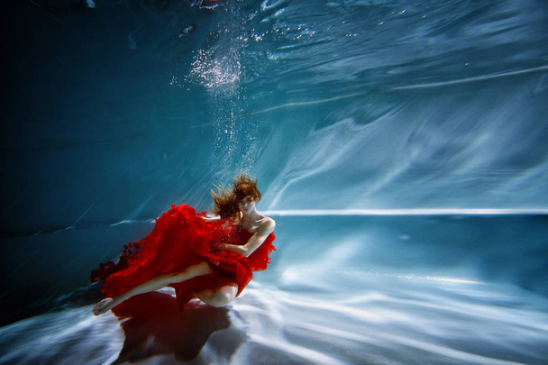 Underwater in the pool with the purest water. Beautiful young girl in a scarlet dress and flowing hair. - Foto, Bild