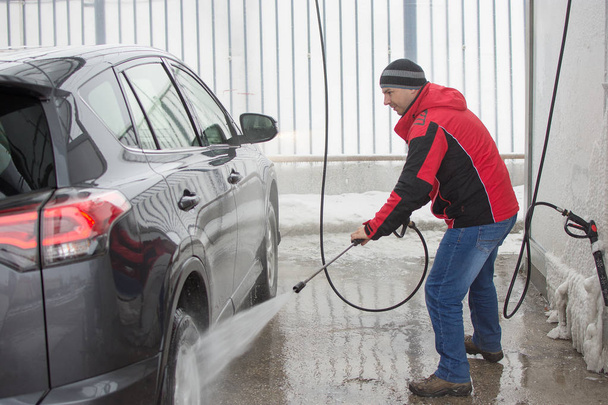 Un hombre con chaqueta roja lavando su coche en la estación de autoservicio con un chorro de alta presión
. - Foto, Imagen