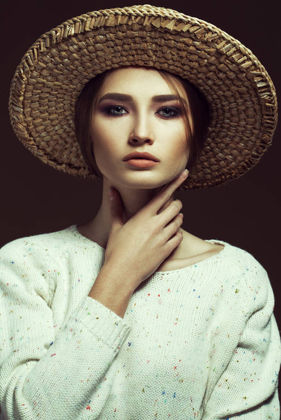 Hipster στυλ. Μοντέρνα πορτρέτο του μια όμορφη κοπέλα φορώντας μοντέρνο λευκό πλεκτό πουλόβερ και ψάθινο καπέλο, θέτοντας πάνω από ρετρό φόντο σοκολάτας. Μόδα στυλ. Studio που γυρίστηκε - Φωτογραφία, εικόνα