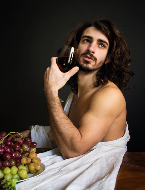 photo half naked curly guy at the table with a plate of grapes and a glass of red drink - Fotografie, Obrázek