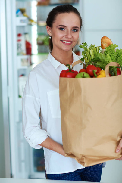 Mujer joven sosteniendo bolsa de la compra de comestibles con verduras. De pie en la cocina - Foto, Imagen
