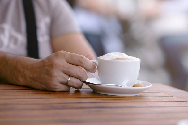 穏やかなリラックス手の姿勢、屋外でのコーヒーのカップを持つカフェで木製のテーブルに若い男の手は、背景をぼかし - 写真・画像
