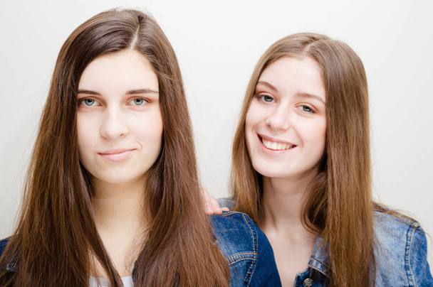 Closeup portrait of two beautiful young women with long dark hair and natural makeup wearing jeans shirts happy smiling looking at camera - Foto, imagen